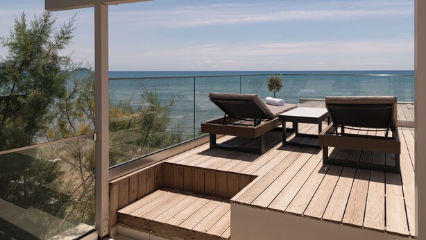 Een houten terras met twee ligstoelen en een kleine tafel, met uitzicht op een rustige zee en verre landschappen.