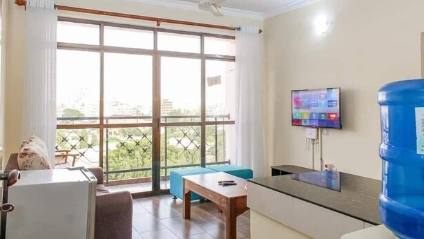 A spacious living area with a large window, a ceiling fan, a flat-screen TV, and a water dispenser. - Lux Suites Juniours Apartment Bamburi (Mombasa)