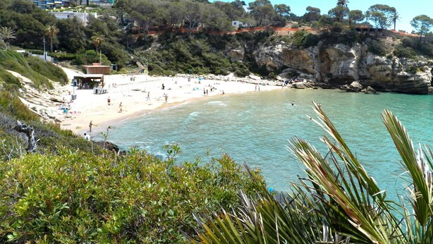 A coastal beach with a sandy shore, a rocky cliff, and buildings in the background. - Idillic penthouse between sea and pine trees (Salou)