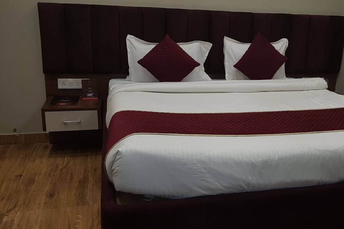 A neatly made bed with white and maroon bedding, a dark headboard, and a bedside table with a lamp and a book.