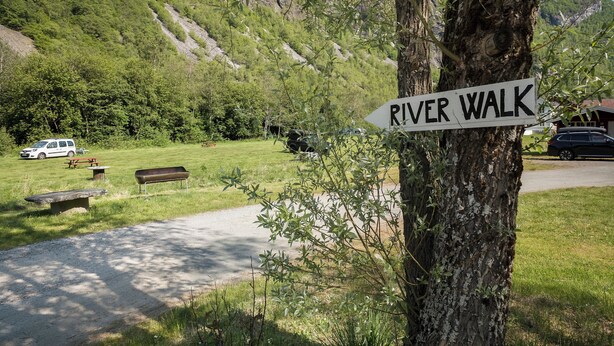 A park with a sign for the River Walk, picnic tables, benches, and a car parked nearby.