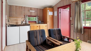 A compact kitchen with a sink, stove, and wooden cabinets. There are two upholstered chairs and a wooden table with a vase of flowers.