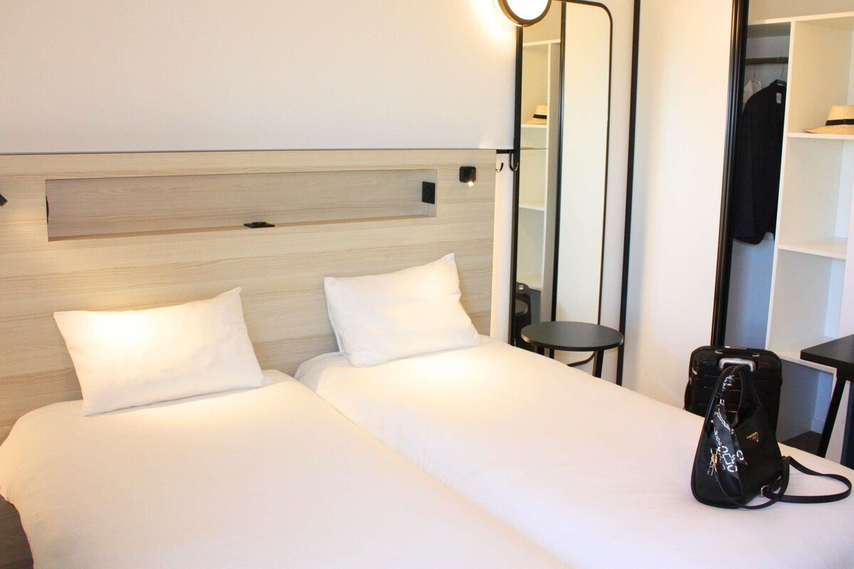 A hotel room with two beds, a bedside table, a mirror, and a wardrobe.
