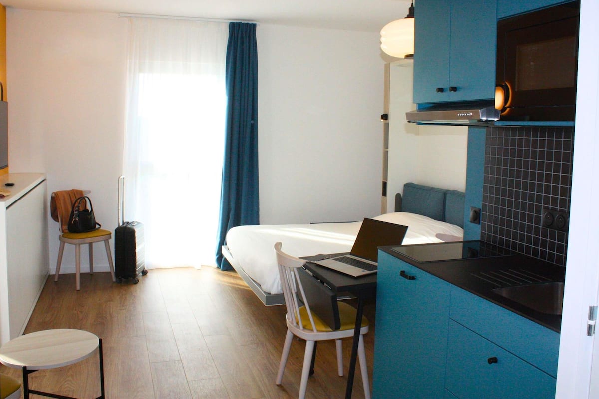 Ein kompaktes Hotelzimmer mit Bett, Schreibtisch mit Laptop, Stuhl, kleinem Tisch, einer Küchenzeile und einem Koffer.