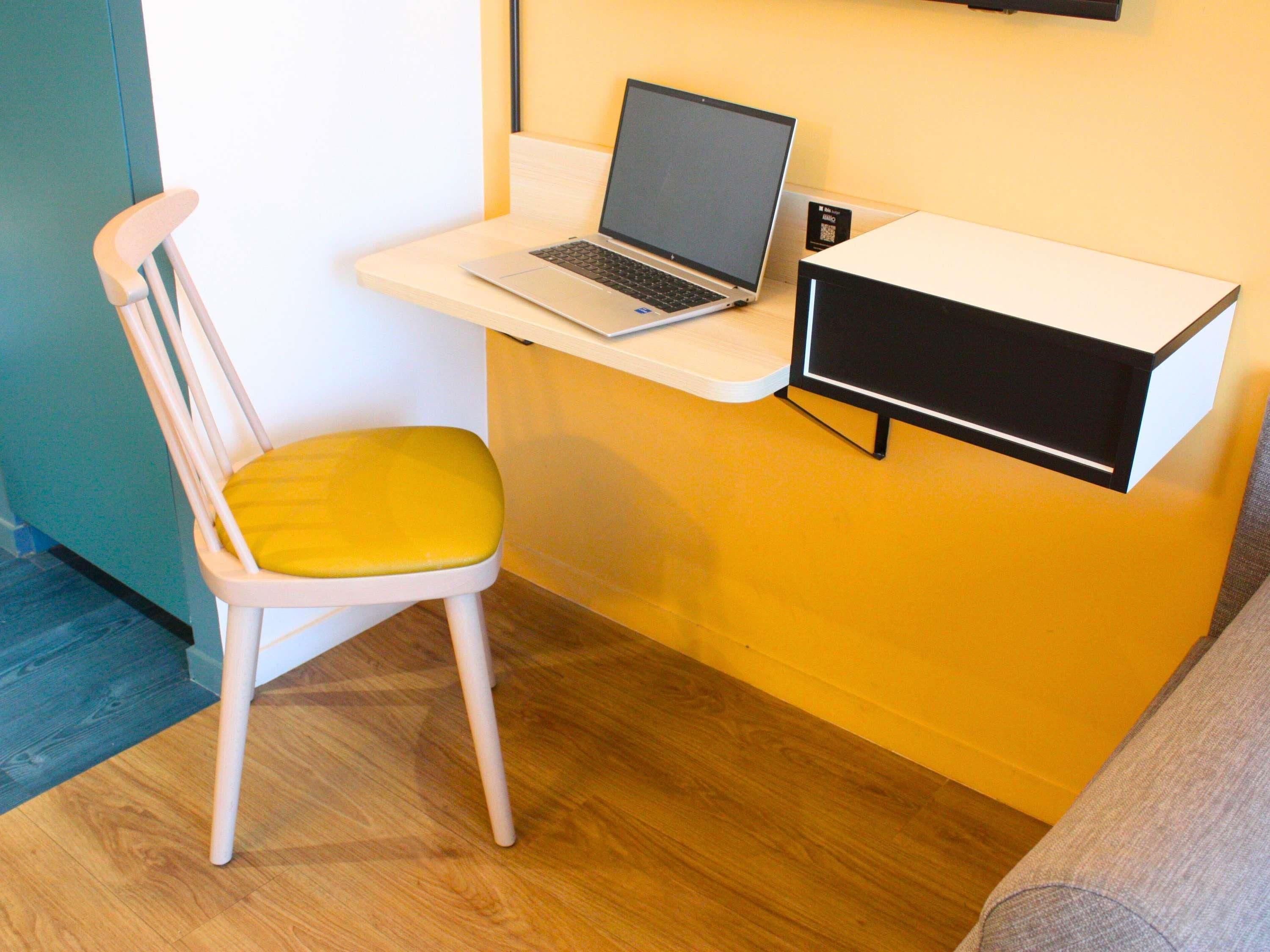 A workspace with a laptop on a white desk, a chair with a yellow cushion, and a black monitor on the wall.