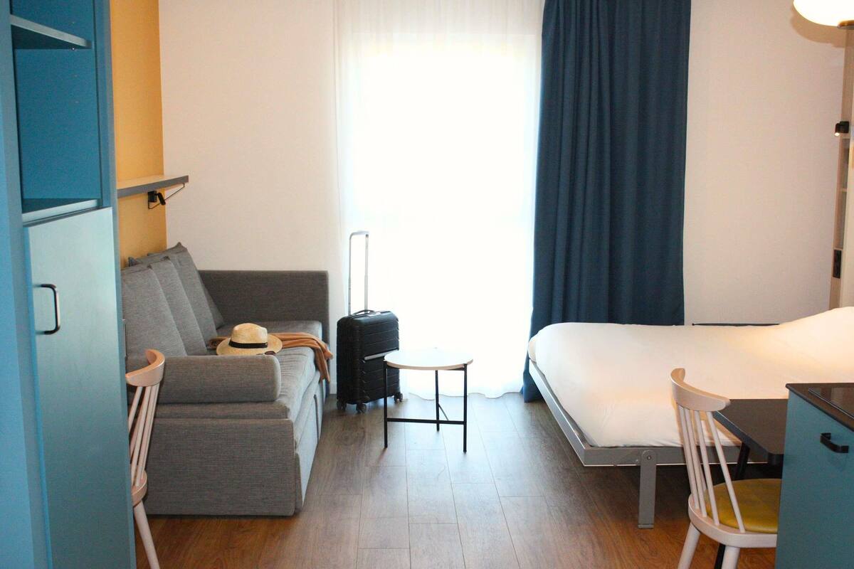 Ein Hotelzimmer mit einem Bett, einem grauen Sofa, einem kleinen Tisch, einem Koffer und einem Stuhl.