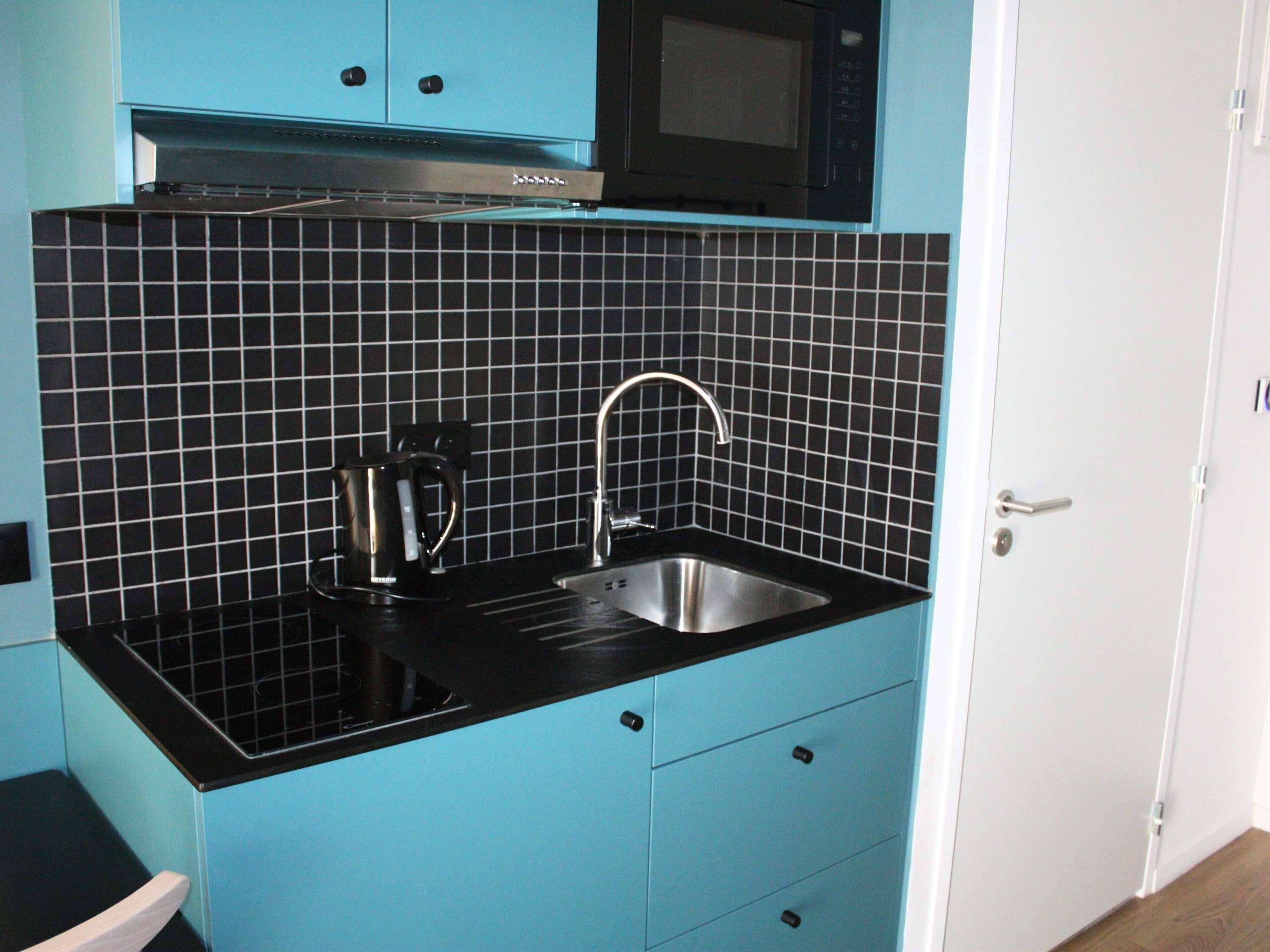 A kitchen with a black countertop, a stainless steel sink, a kettle, and a microwave.