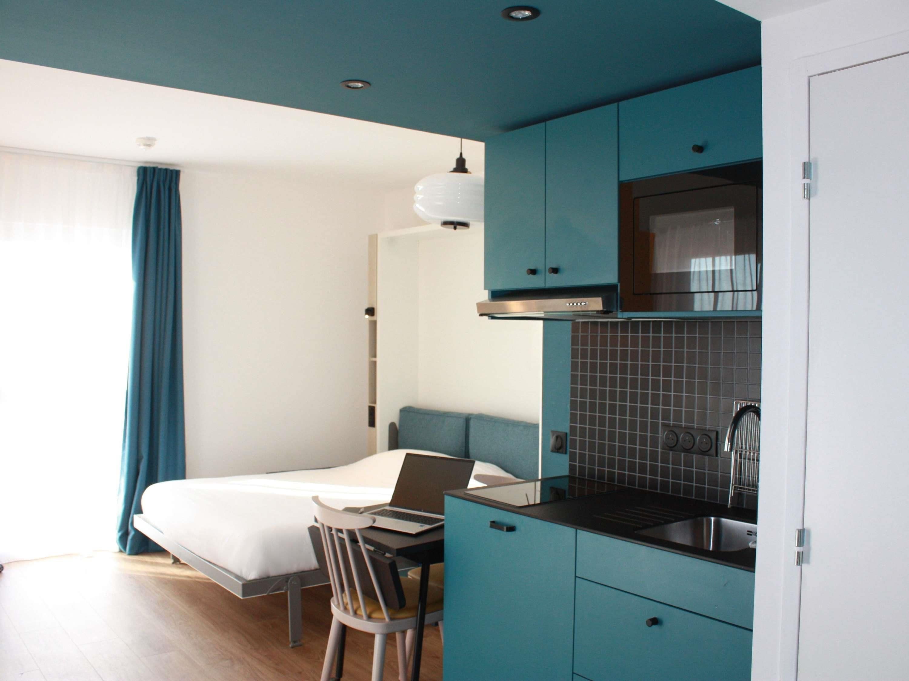 A compact room with a bed, a desk with a laptop, a kitchenette, and a window with curtains.