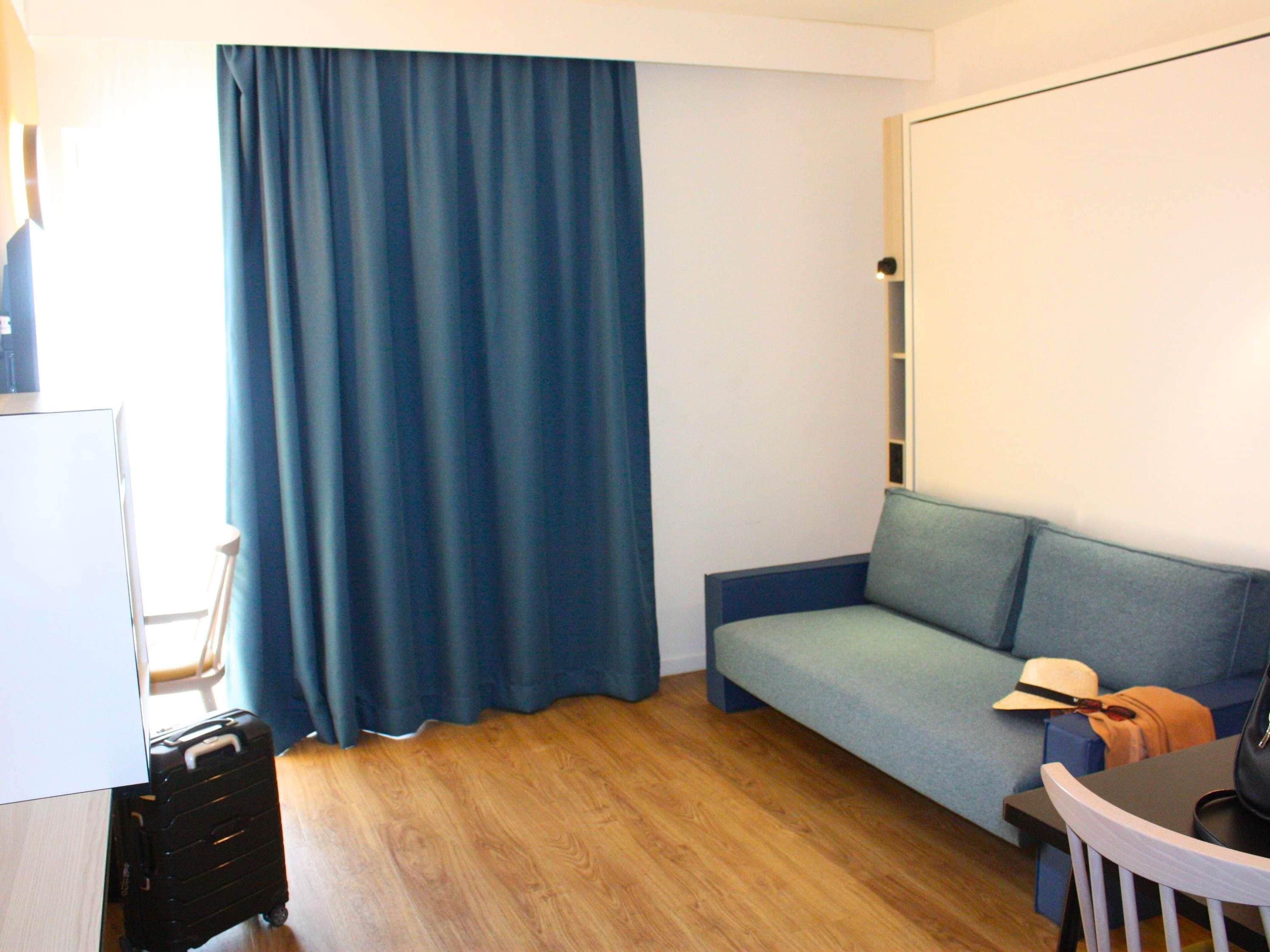 A room with a wooden floor, a blue curtain, a grey sofa, a small table with a hat and a bag, and a whiteboard on the wall.