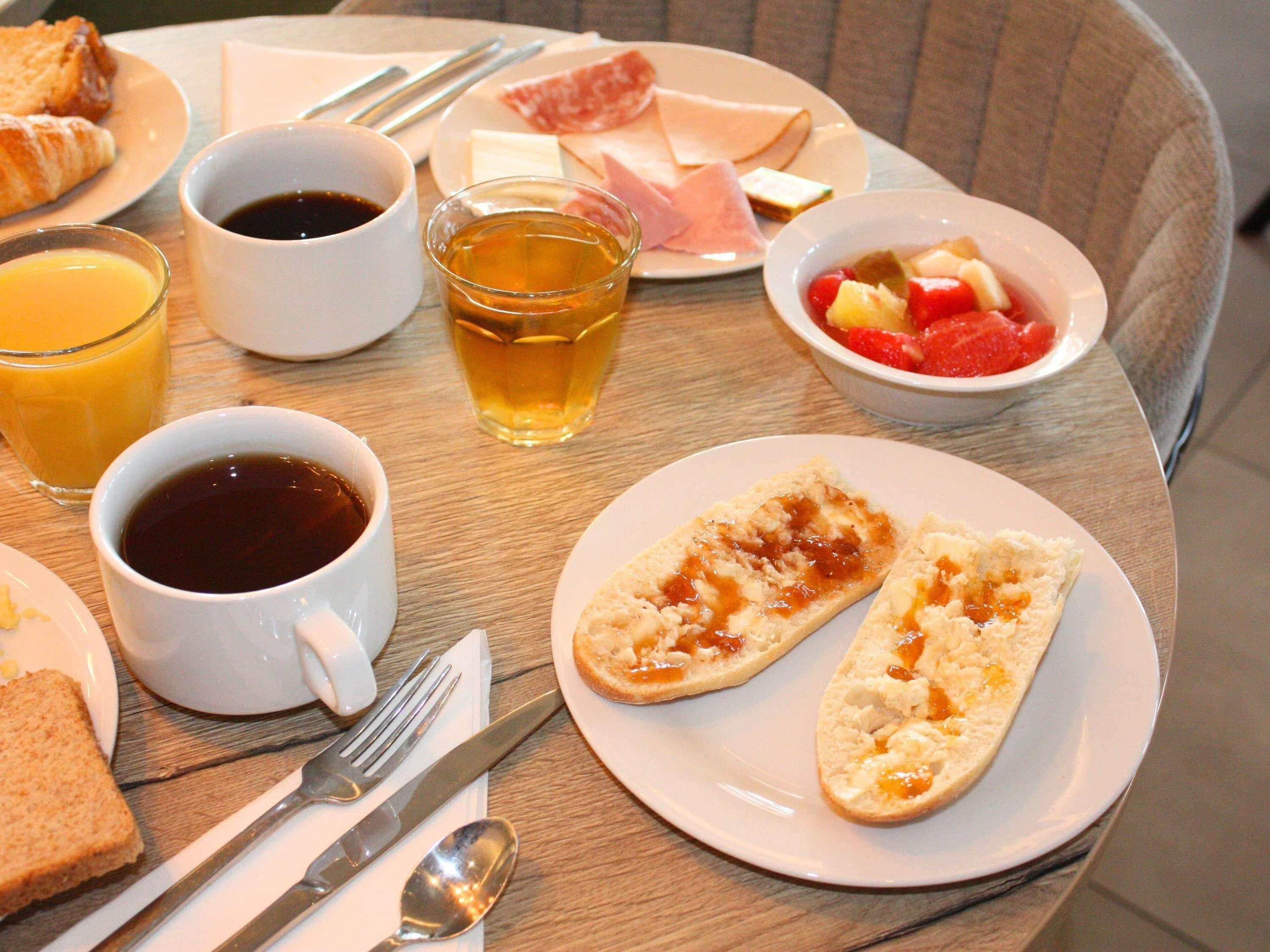 Impostazione colazione con croissant, prosciutto, formaggio, frutta e bevande su un tavolo.