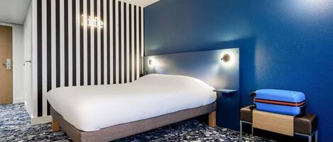 Ein Hotelzimmer mit einem Bett, einem an Beleuchtung versehenen Kopfteil, einer blauen Wand, einem gestreiften Raumteiler und einem Gepäckwagen.