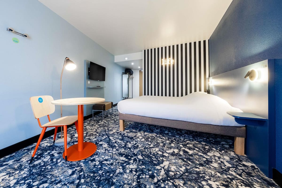 Ein modernes Hotelzimmer mit Bett, Schreibtisch, Stuhl, Fernseher und Wandlampe.