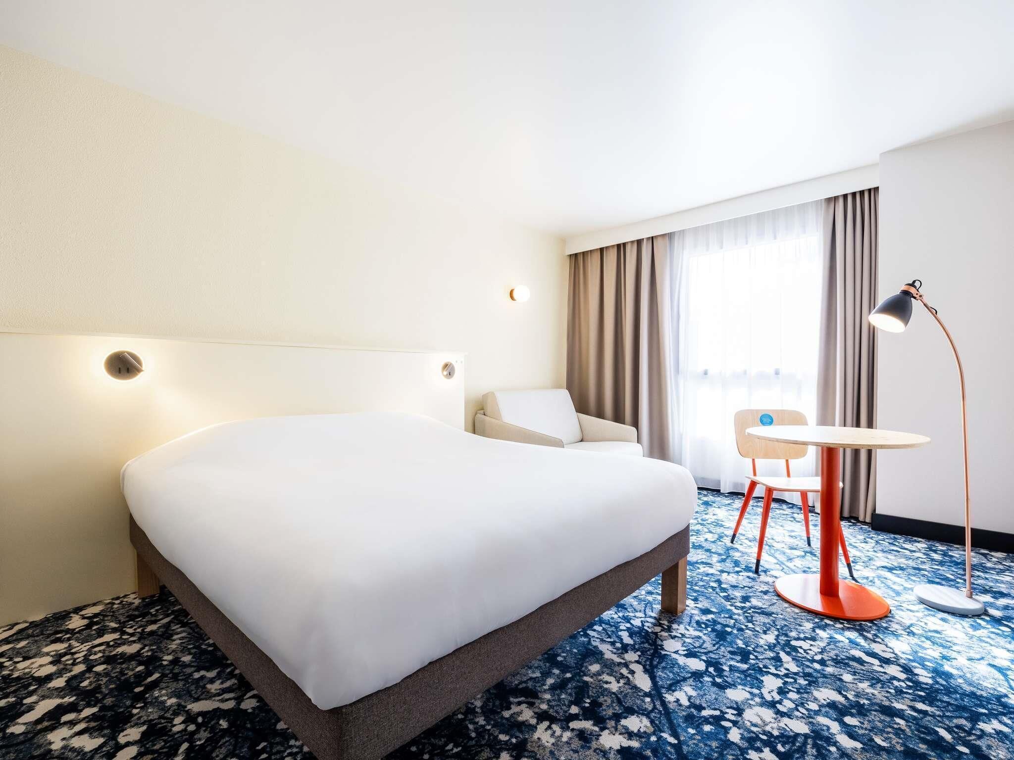 Habitación de hotel con cama, una silla, una mesita, una lámpara y vista a la ciudad por la ventana.