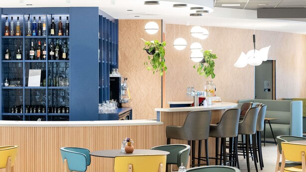 Een moderne bar met diverse drankflessen, een koffieapparaat en een zithoek met ronde tafels en stoelen.