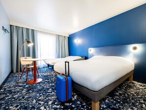 Une chambre d’hôtel avec un lit, un bureau, une chaise, une valise et une fenêtre avec des rideaux.
