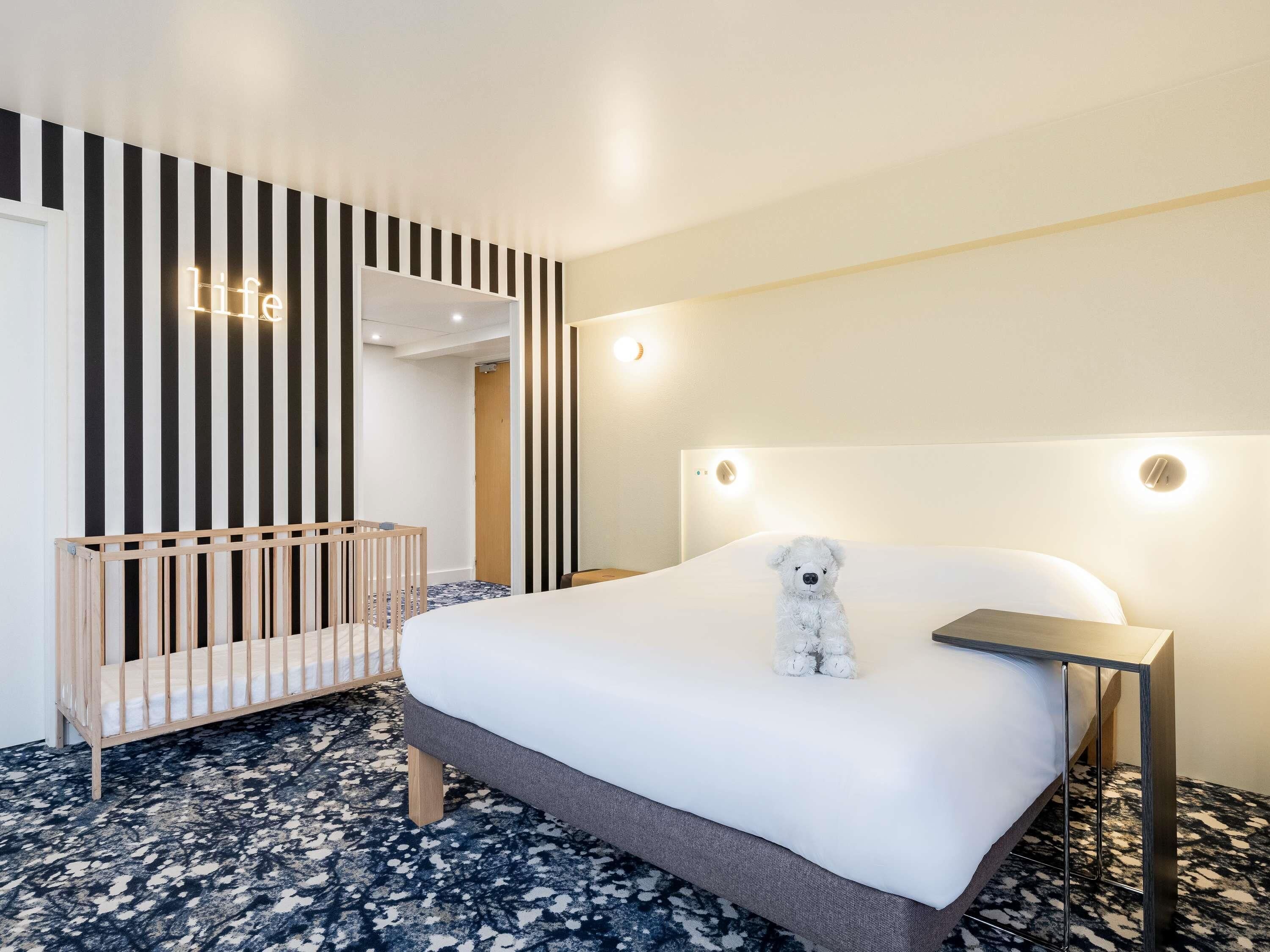 Une chambre d’hôtel avec un lit, un berceau, un ours en peluche et un mur à rayures verticales.