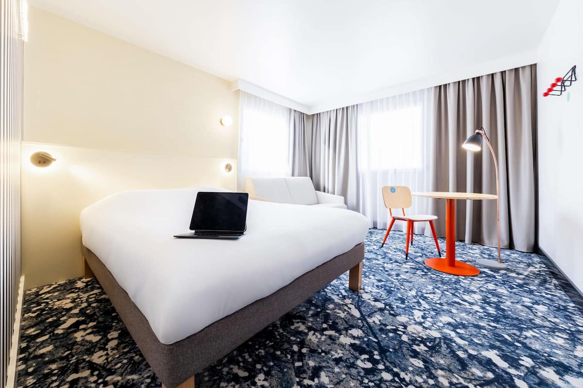 Ein Hotelzimmer mit einem großen Bett, einem Schreibtisch mit Stuhl, einem Laptop und einer Schreibtischlampe.