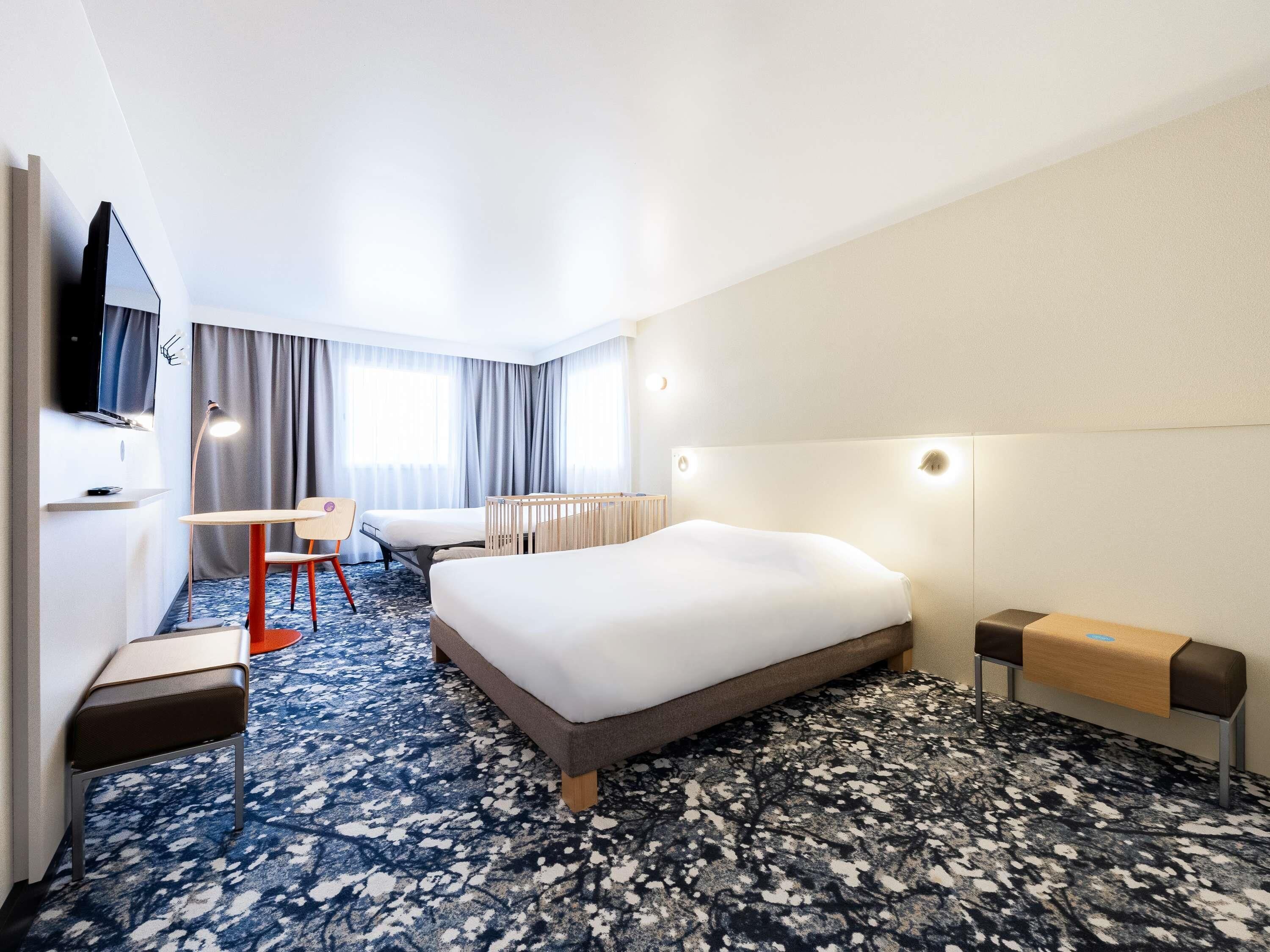 A hotel room with a large bed, a desk, a chair, a TV, and a crib.