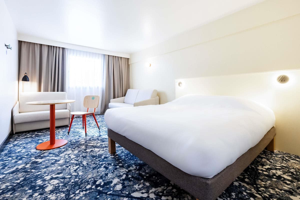 Ein Hotelzimmer mit einem großen Bett, einem kleinen Tisch, einem Stuhl, einer Couch und einem Fenster mit Vorhängen.