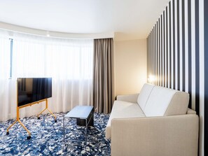 Ein Hotelzimmer mit einer Couch, einem Fernseher auf einem Schrank, einem kleinen Tisch und einem gemusterten Teppich.