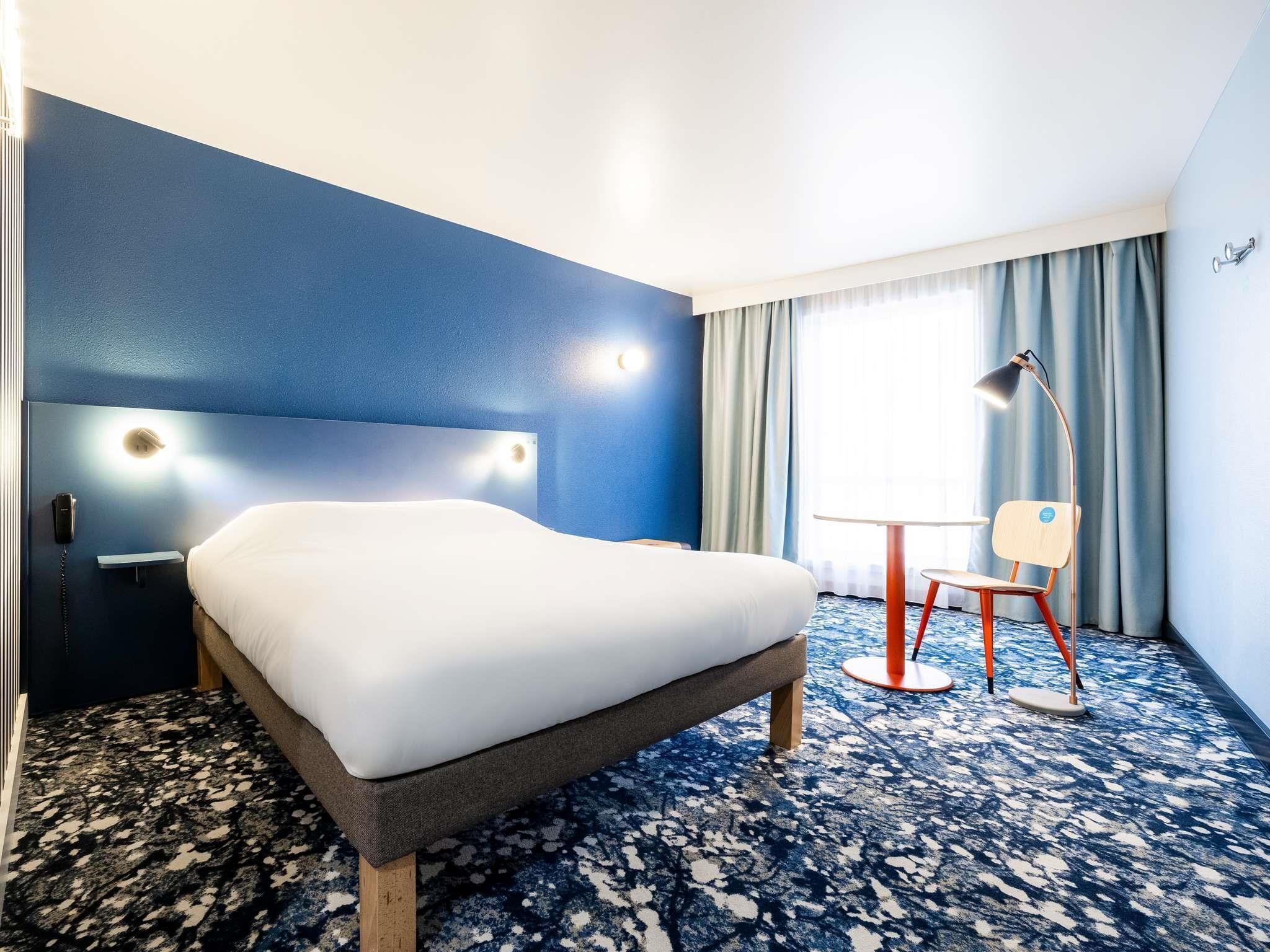 Habitació d'hotel amb un llit, una taula, una cadira, una làmpada i una finestra amb cortines.