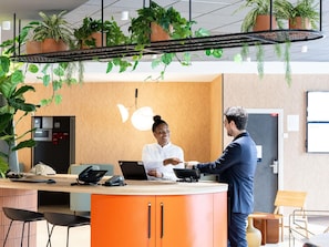 Een receptiebalie met een zittende vrouw, een staande man en hangende plantenpotten erboven.