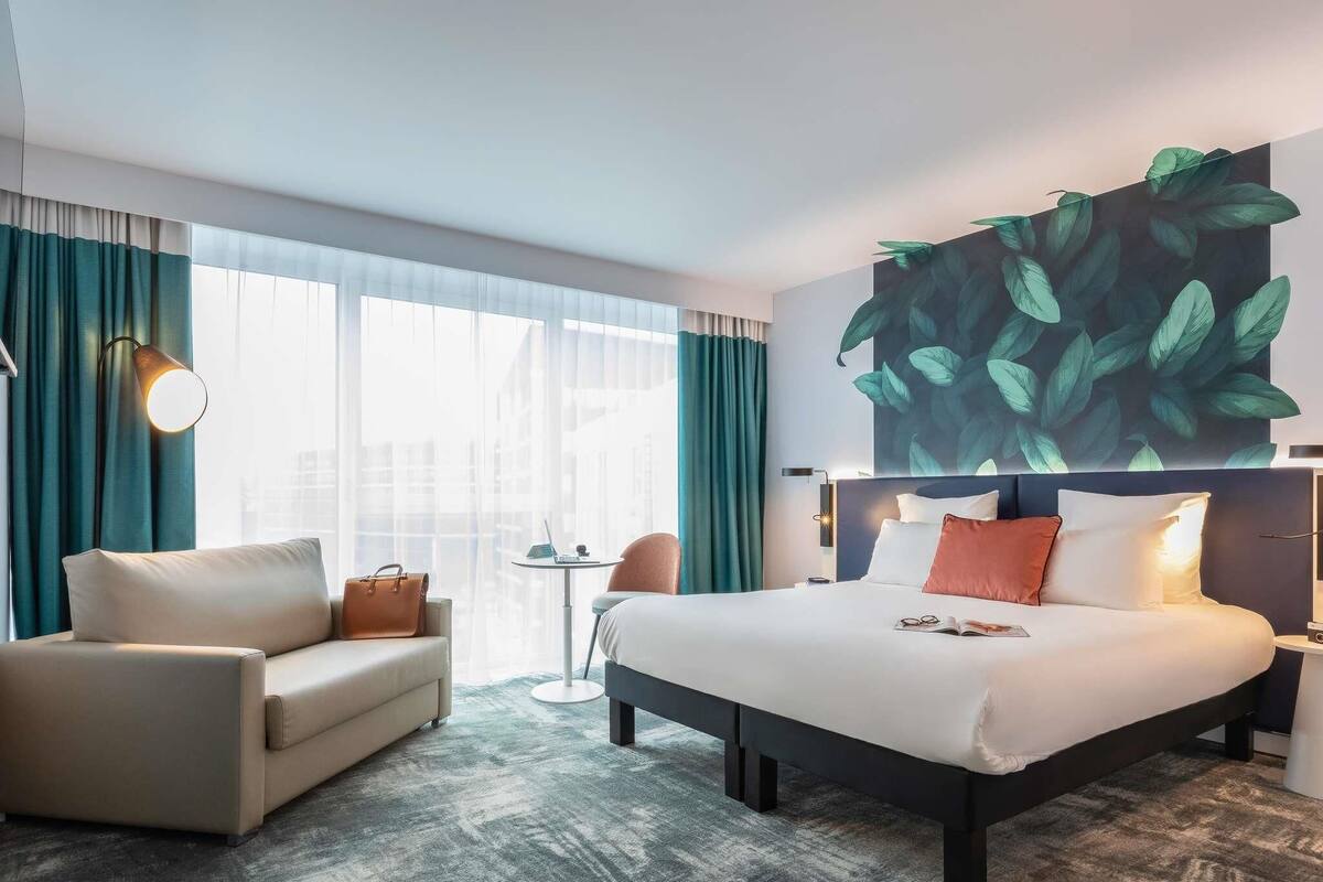Ein Hotelzimmer mit einem Bett, einer Couch, einem kleinen Tisch und einem großen Fenster mit Vorhängen.
