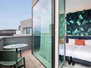 A modern hotel room with a city view, a balcony with green chairs, and a large leafy wall mural.