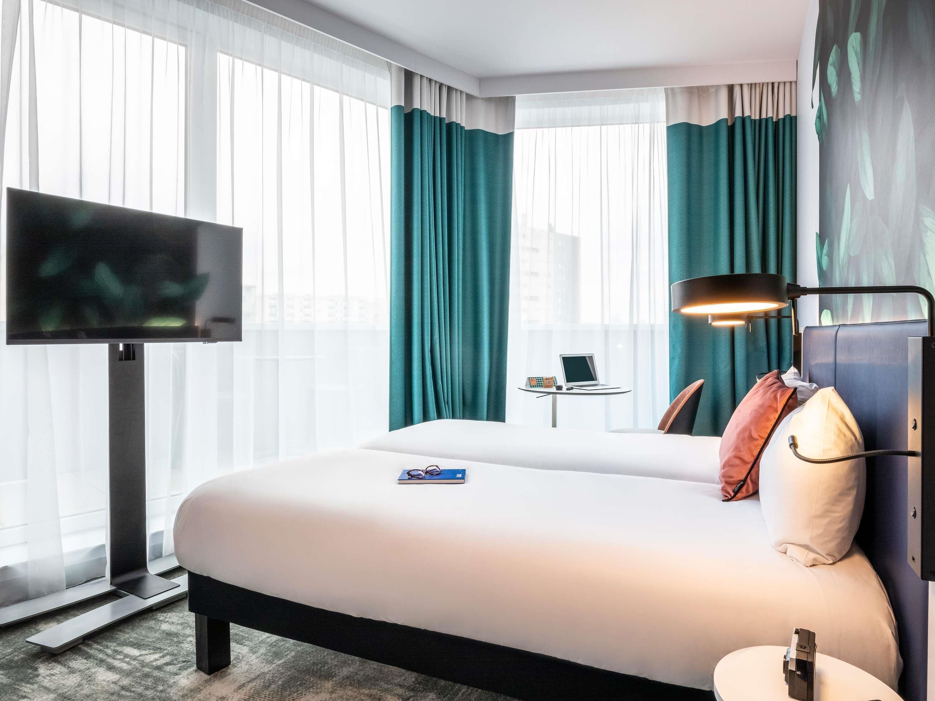 A hotel room with a bed, a TV, a desk with a laptop, and a window with curtains.