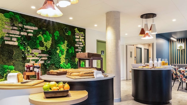 A modern hotel lobby with a green wall, wooden furniture, and a breakfast bar with pastries and fruits. - Ibis Styles Bezons Paris La Défense (Opening March 2024) (Bezons)