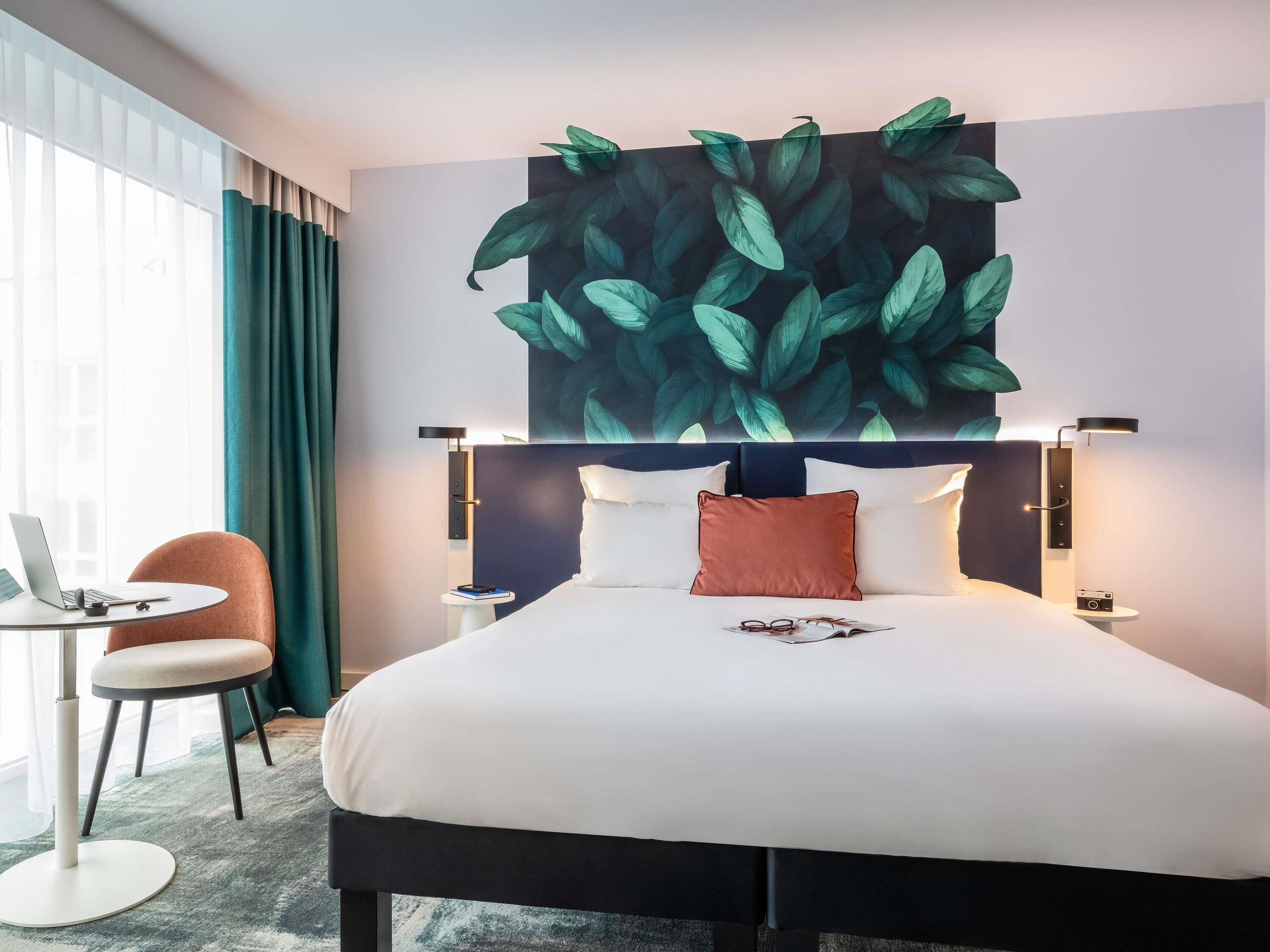 A hotel room with a bed, two bedside lamps, a desk with a laptop, a chair, and a large wall mural.