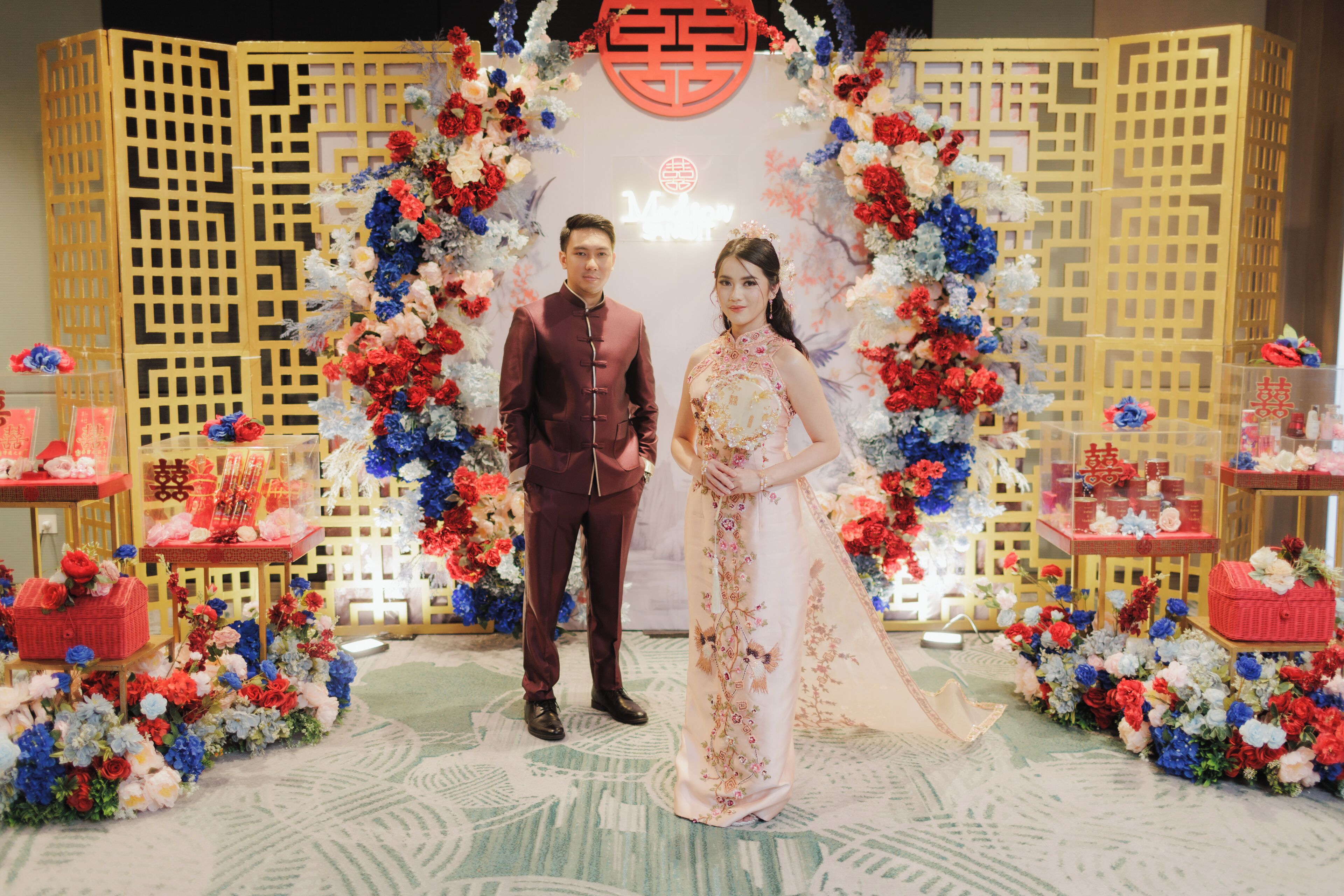 A couple standing in front of a decorative backdrop with floral arrangements and traditional Chinese symbols.
