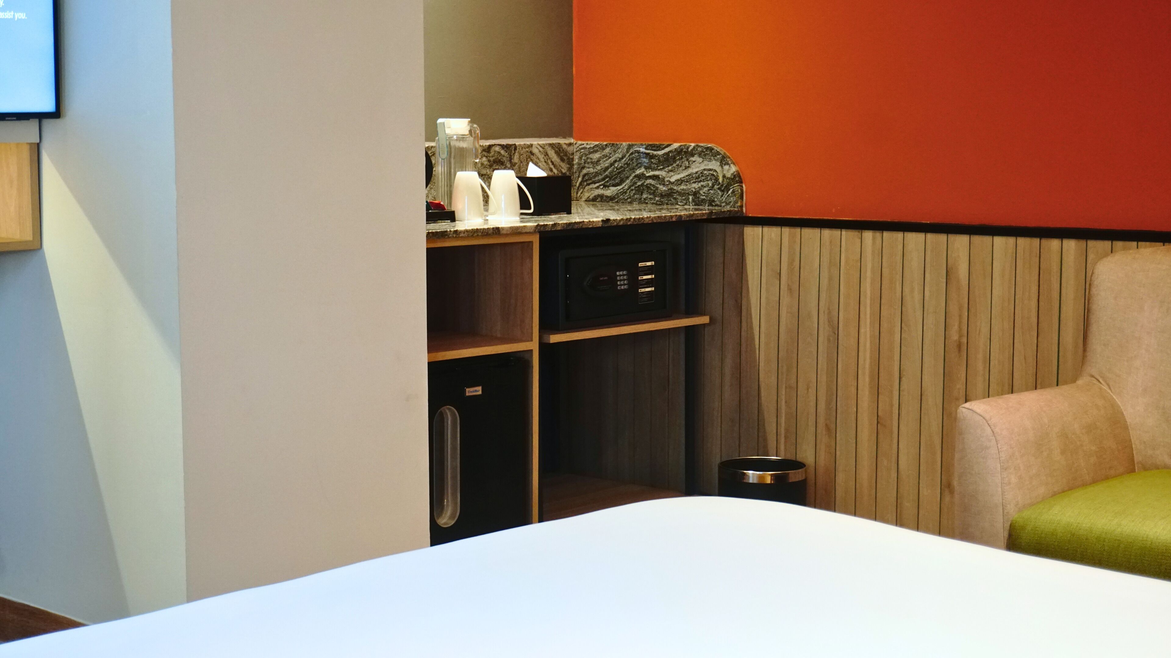 A hotel room with a bed, a small kitchenette with a microwave, a trash bin, and a chair.