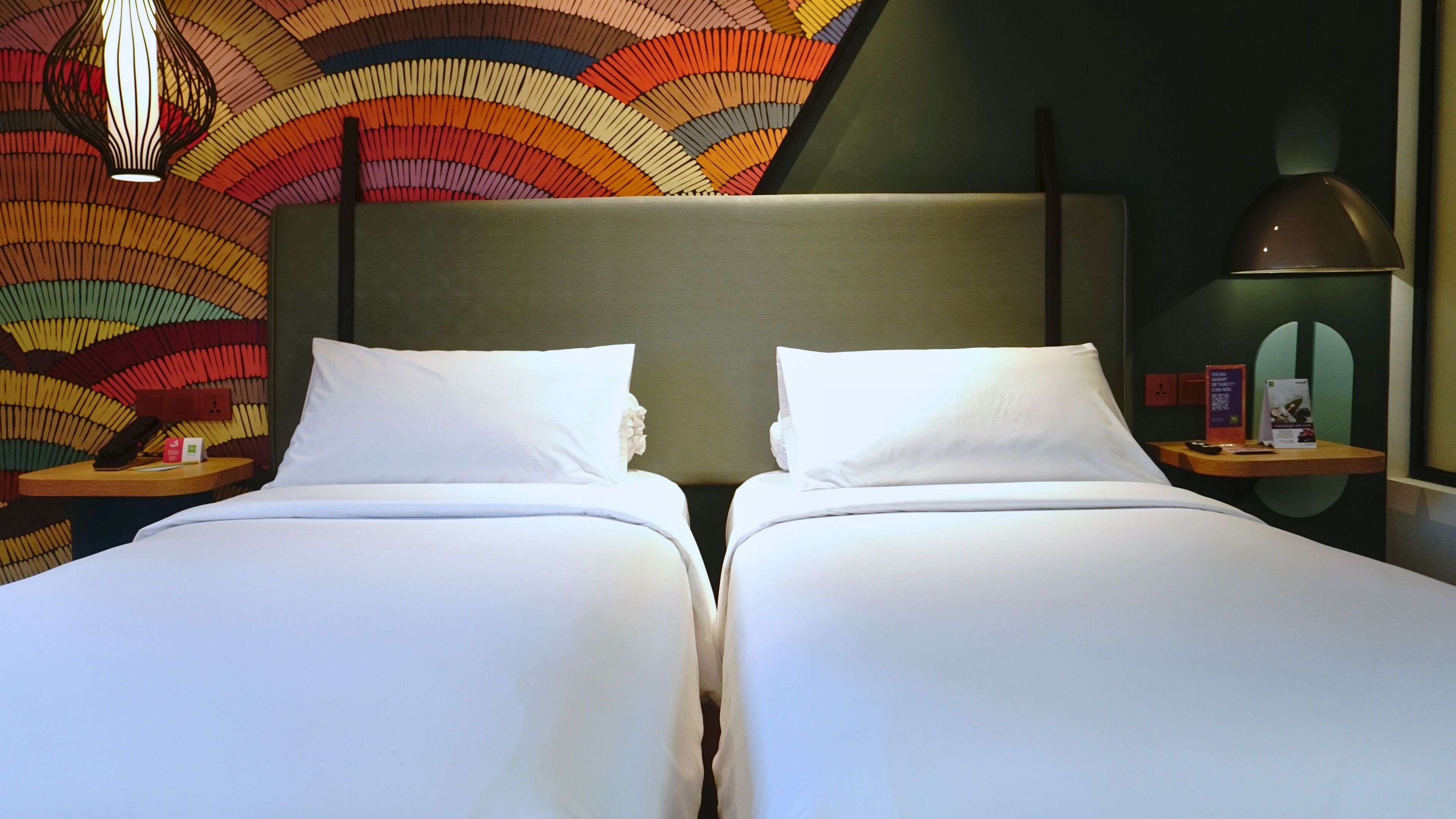 A hotel room with two beds, a patterned wall, a modern lamp, and a small table with a phone and a brochure.