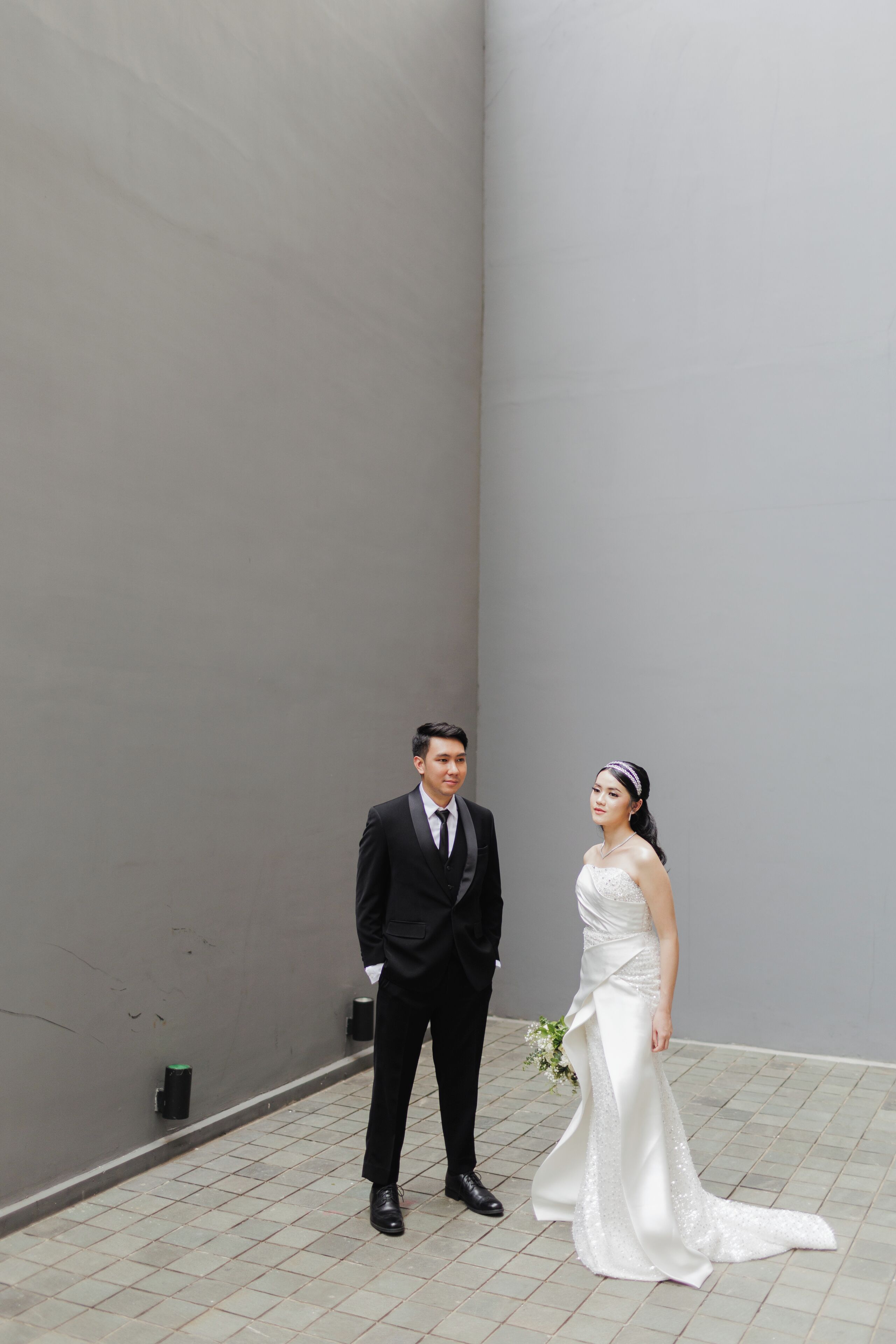 A man and woman in wedding attire standing in a narrow corridor with a plain wall and tiled floor.