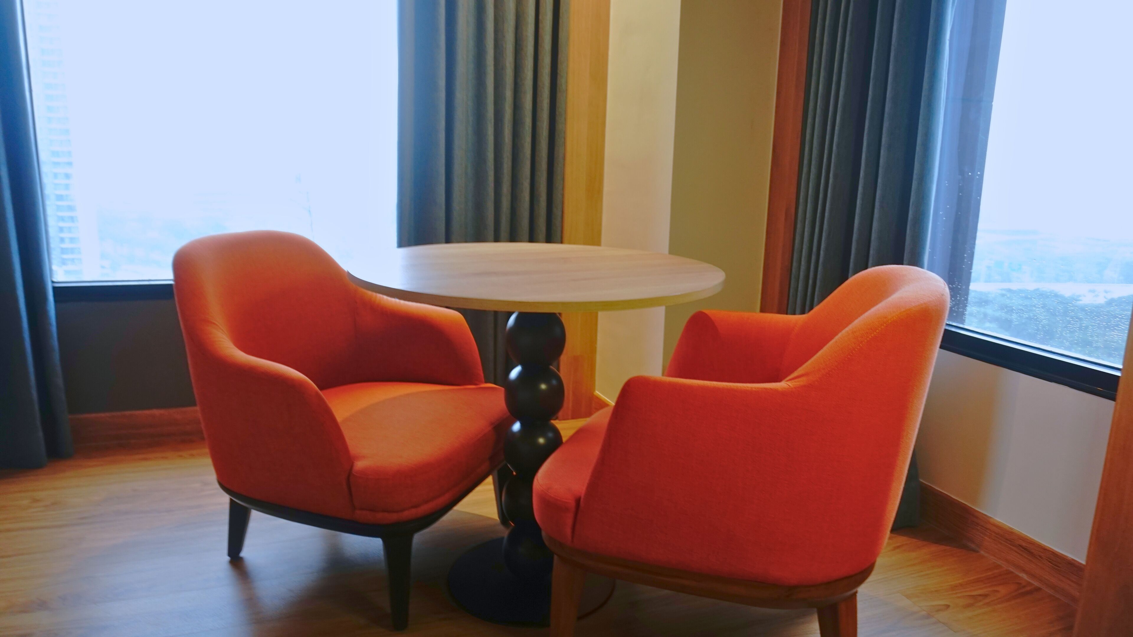 A room with two orange chairs around a small round table, large windows with curtains, and a view of a cityscape.