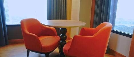 A room with two orange chairs around a small round table, large windows with curtains, and a view of a cityscape.
