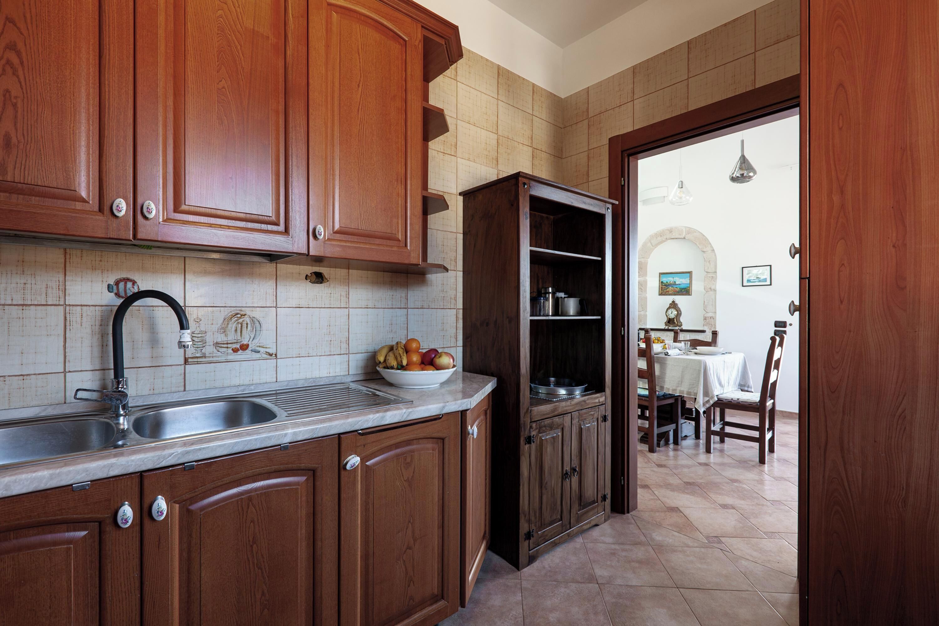 A kitchen with wooden cabinets, a marble countertop, and a dining area visible through an open door.