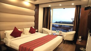Una habitación de hotel con una cama grande, dos almohadas rojas, una mesita de noche con un control remoto, una silla y vistas a la ciudad desde la ventana.