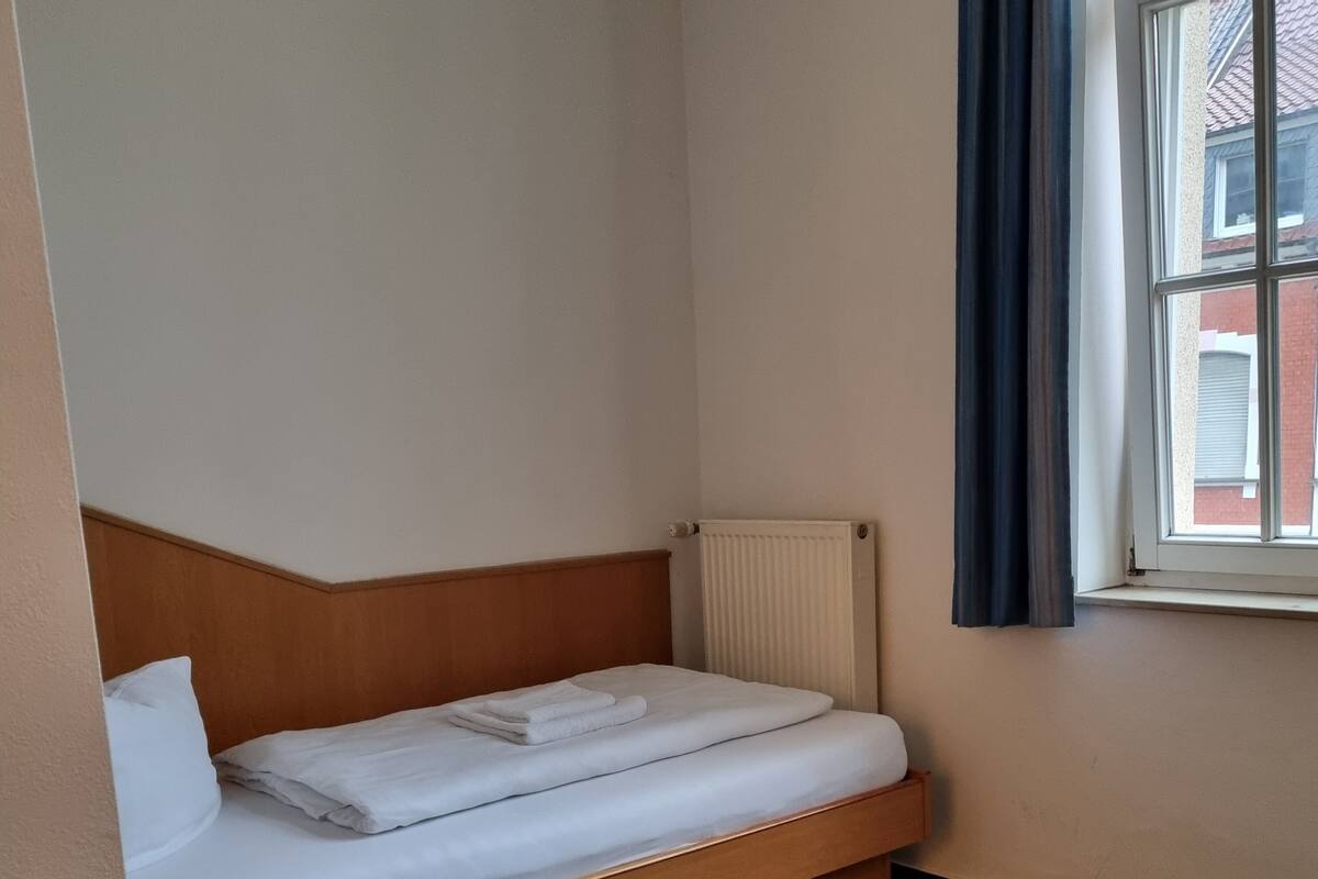 A hotel room with a single bed, a wooden nightstand, a radiator, and a window with curtains.