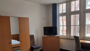 A hotel room with a bed, a TV, a chair, a wardrobe, and a window with a view of buildings. - Stadthotel im Kolpinghaus  (Werne)