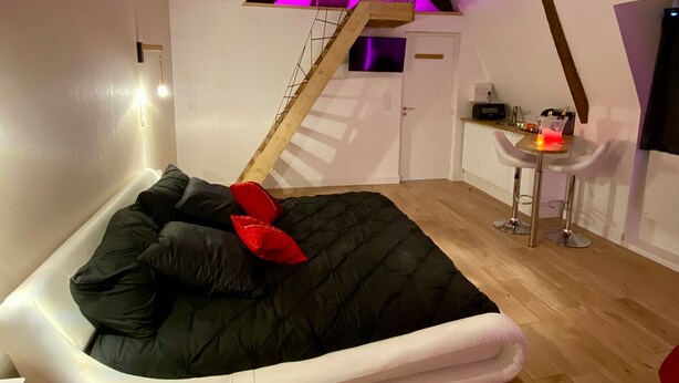 Ein Schlafzimmer im Loft-Stil mit schrägem Dach, einem Bett mit schwarzen und roten Kissen, einer kleinen Küchenzeile und einer Treppe.