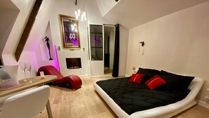 Ein modernes Schlafzimmer mit einem großen Bett, einem Schreibtisch mit Stuhl, einem Kamin und Blick ins Freie durch eine Glastür.