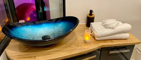 Ein modernes Badezimmer mit einem schwarzen und blauen Glastischwaschbecken, einer kleinen, beleuchteten Kerze und ordentlich gefalteten weißen Handtüchern auf einer Holzkommode.