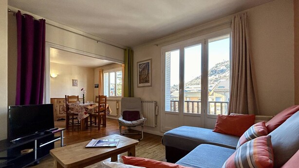 A living room with a sofa, a TV, a dining table with chairs, and a window with a view of buildings and hills.