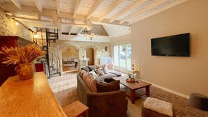 A spacious living area with a high ceiling, a stone wall, a wooden staircase, and a TV mounted on the wall. - Charming Cottage with Private Heated Pool, Fitness Room, Game Room, Near Rillé Lake (Rillé)
