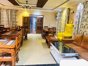 Un vestíbulo de hotel con comedor, un sillón amarillo, un sofá marrón y un televisor sobre un soporte blanco.