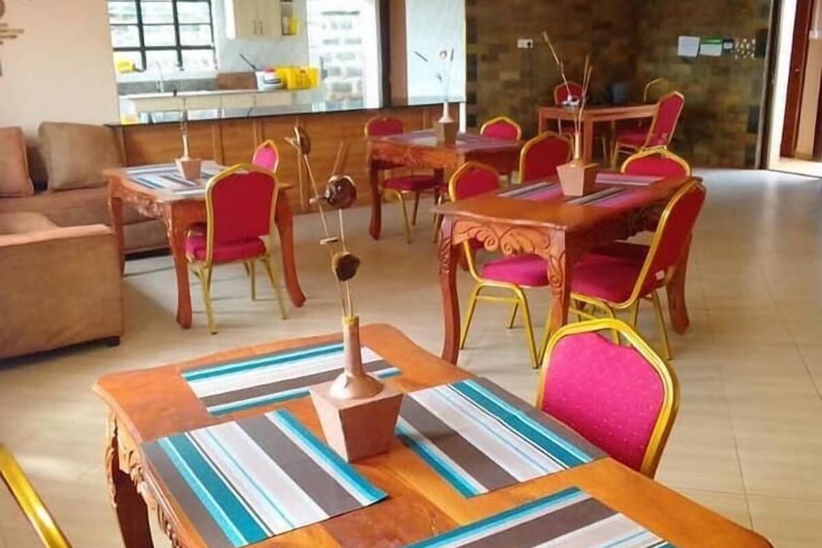 A dining area with wooden tables and red chairs, a kitchen in the background, and a large window.