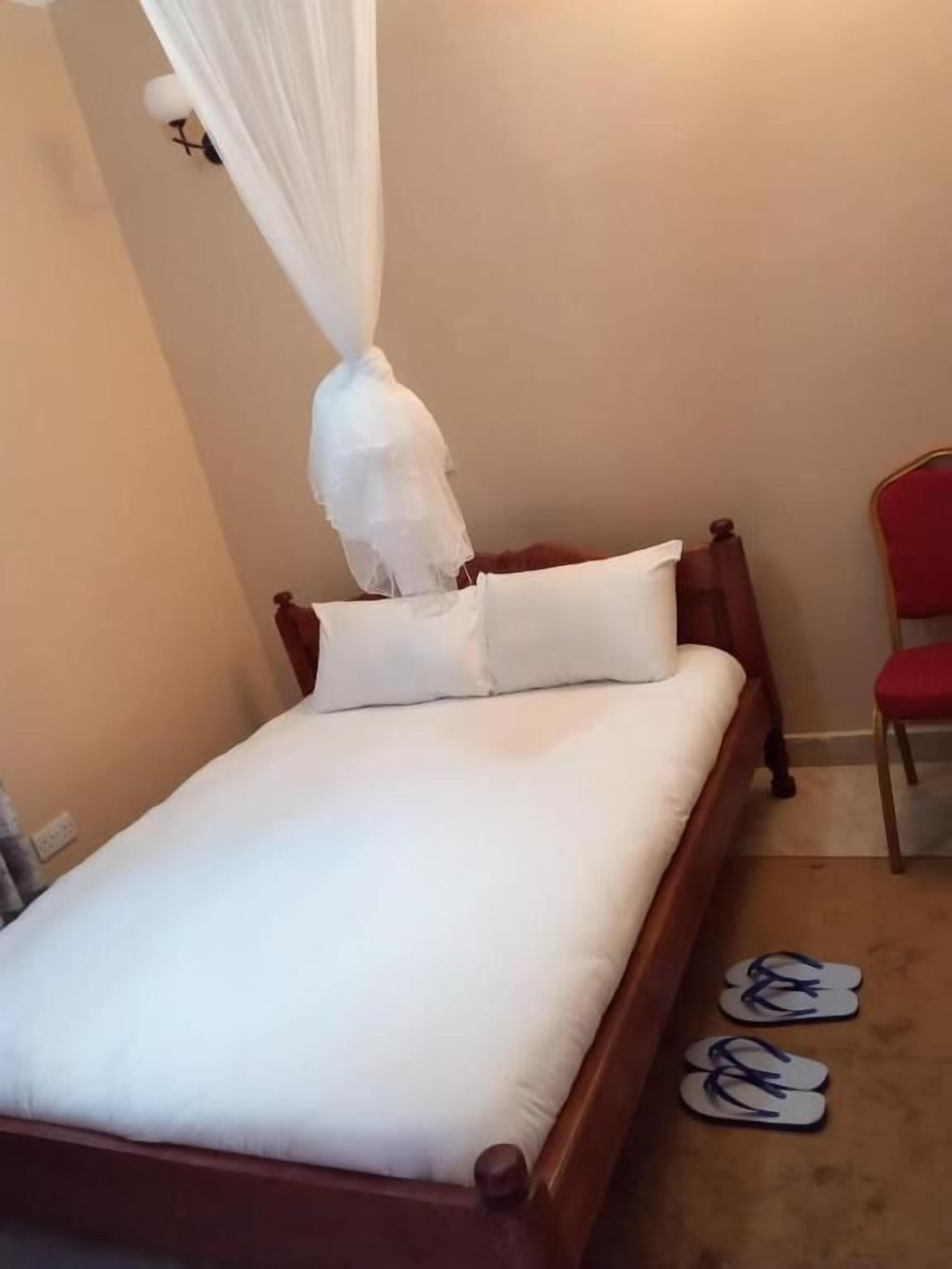 A neatly made bed with white bedding and a canopy, a wooden headboard, and a pair of flip-flops on the floor.