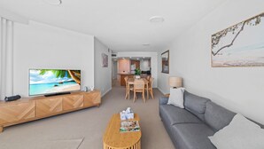 A modern living room with a flat-screen TV, a grey sofa, a wooden coffee table, and a dining area with a wooden table and chairs. - Zanzibar 505 amazing beachfront resort apartment withm pool , spa and access to gym (Mooloolaba)